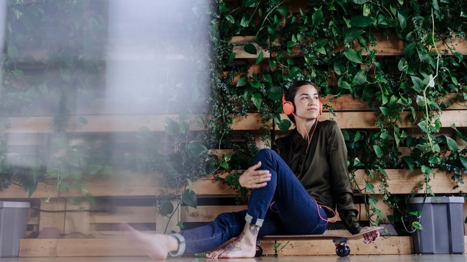 Lady sitting on her skateboard whilst listening to music, surrounded by green leaves and plants